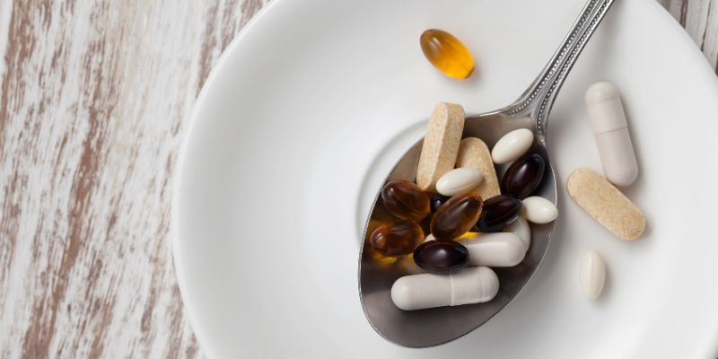 A spoon full of various pills and supplements on a white plate with a wooden table background.