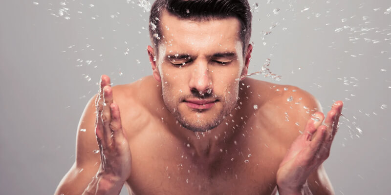 A shirtless man claps his hands together, causing water droplets with face wash to splash around him against a light grey background.