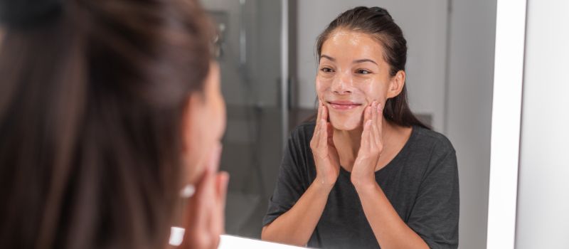 A woman in a gray t-shirt applying facewash product to her cheeks while standing in front of a bathroom mirror.