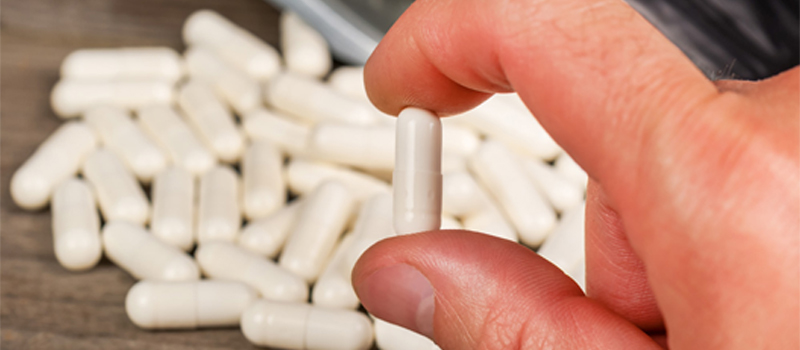 A person holds a single pill in front of a large pile of various pills, highlighting Gluta White Capsules.