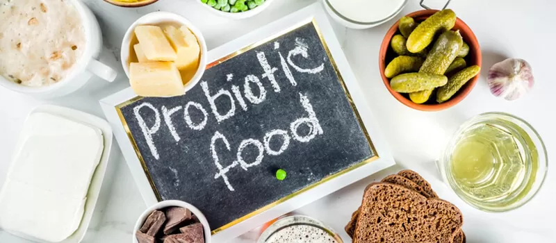 A variety of probiotic foods including cheese, pickles, fermented foods, and bread are displayed around a chalkboard labeled "Probiotic Food