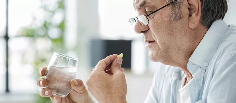 An older man holds a glass of water while taking a pill, contemplating the daily use of Ginglitz Capsules.