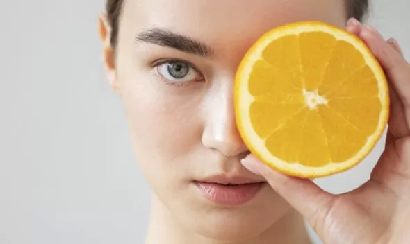 A woman presents an orange slice to her face, highlighting the importance of antioxidants in skincare routines.