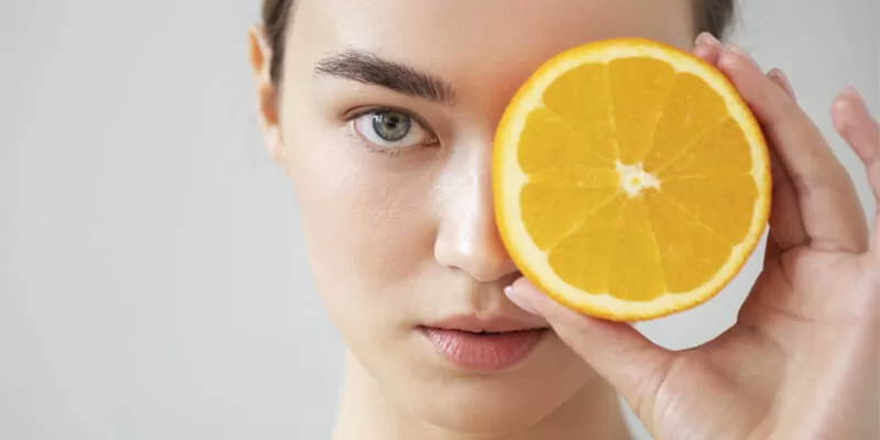 A woman presents an orange slice to her face, highlighting the importance of antioxidants in skincare routines.